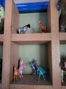 A wooden shelf showing two groups of miniature model horses. One shelf has two, a blue roan and a chestnut horse. The other has four unicorns in gold, pink, purple and light blue.